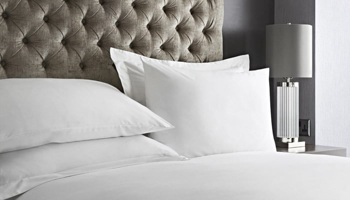 A hotel bed dressed in crisp white Prima bed linen with a patterned cushion